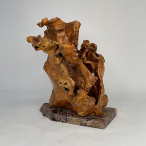 Może przedstawiać: Unikalna rzeźba z drewna o naturalnej, organicznej formie. Rzeźba ma ciepły brązowy kolor i charakteryzuje się skomplikowanymi detalami i fakturami. Jest zamontowana na prostokątnej podstawie z nakrapianym fioletowo-szarym wzorem.