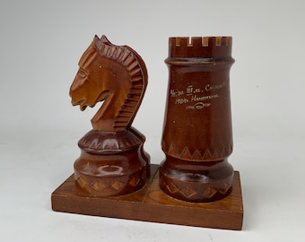 May include: A brown wooden chess set piece holder featuring a carved knight and a rook on a rectangular base. The rook has engraved Cyrillic text that reads "Уз за М. Стрел" and "1984г. Никопол". Both pieces have geometric diamond patterns carved around their bases.