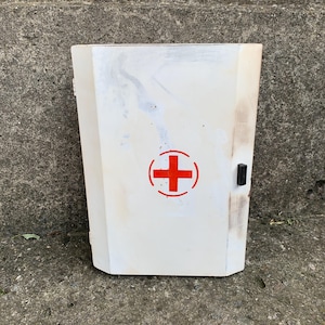 May include: A rectangular white wall-mounted cabinet featuring a red cross symbol centred on the front door. The cabinet has a small black latch on the right side and shows signs of wear, including surface scuffs and discolouration on the finish.