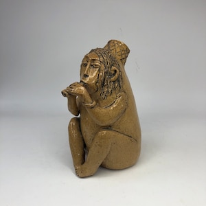 May include: A ceramic sculpture of a person playing a flute. The sculpture is beige with brown details and has a basket on its head.