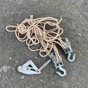 May include: A set of vintage rigging equipment, including two grey pulleys with hooks, a rope, and a silver rope clamp. The rope is beige and coiled. The items are arranged on a textured grey surface.