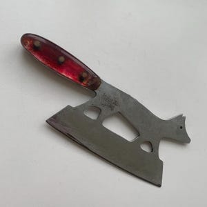 May include: A vintage multi-tool with a red handle and a stainless steel blade. The blade has a unique shape with cutouts and a fox-like design at the end. The handle is secured with three rivets.
