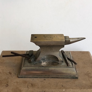 May include: A miniature metal anvil with a pointed horn, set on a rectangular base with two small metal rods. The anvil is made of brass and silver.