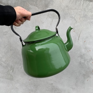 Puede incluir: Una tetera de esmalte verde con asa y boquilla negras. La tetera tiene tapa y la sostiene una persona. Un artículo de menaje de cocina clásico.