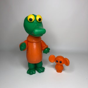 Op de afbeelding: Een groene krokodil met een oranje trui staat naast een kleine oranje muis met grote oren en blauwe ogen. Beide figuren zijn van plastic.