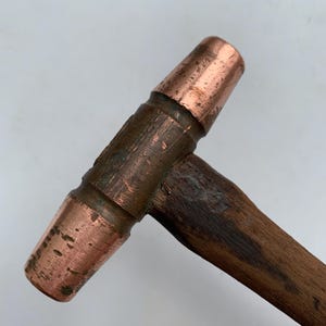 Puede incluir: Un martillo con cabeza de cobre y mango de madera. La cabeza del martillo es de color marrón rojizo y el mango es de color marrón oscuro. El martillo está diseñado para golpear y dar forma a los materiales.