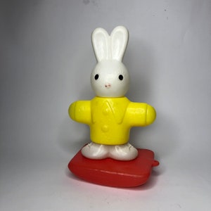 May include: A white bunny wearing a yellow jacket is standing on a red rocking base. The bunny has black eyes and a pink nose.