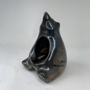 Könnte beinhalten: Eine glänzende, dunkelmetallische Keramikskulptur in Form eines sitzenden Bären mit einer ausgehöhlten Mitte. Die Oberfläche weist ein reflektierendes, irisierendes Finish mit tiefen Anthrazit- und Bronzetönen auf und zeigt eine glatte, runde Form.