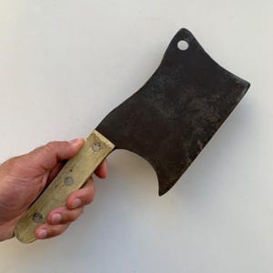 May include: A vintage cleaver with a dark, aged blade and a light-colored wooden handle. The cleaver has a curved edge and a hole near the top. The handle is attached with three rivets. The cleaver is designed for chopping and cutting.