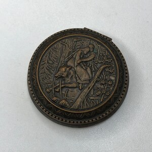 May include: A round, bronze-colored compact mirror with an intricate design. The mirror features a raised image of a woman riding a wolf through a forest.