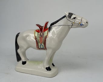 May include: A white ceramic horse figurine with a black mane and tail, and a colorful saddle. The saddle is red, yellow, and green. The horse is standing on a rectangular base.