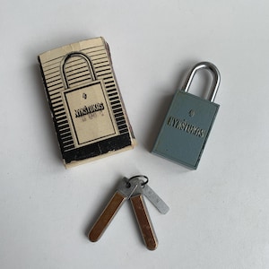 May include: A vintage padlock with a blue metal body and a silver shackle. The padlock has the word "NYKŠTUKAS" printed on it. The padlock is accompanied by a key and a small cardboard box with the same branding.