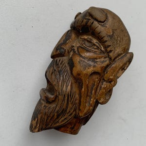 Op de afbeelding: Een gebeeldhouwde houten sculptuur van het gezicht van een bebaarde man in profiel. De sculptuur heeft een warme bruine kleur met gedetailleerde gravures van het haar, de baard en de gelaatstrekken. Het hout heeft zichtbare nerven en wat slijtage.