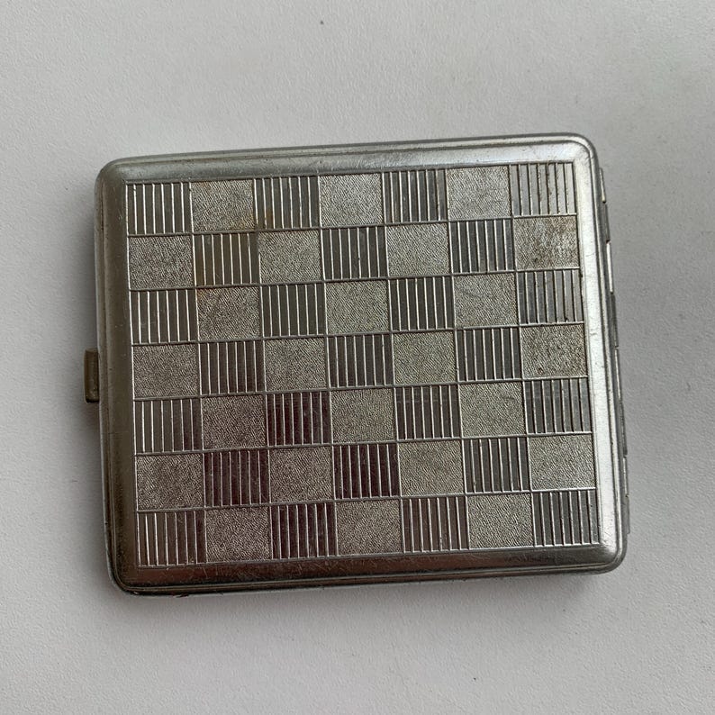 May include: A silver metal cigarette case with a chequered pattern.