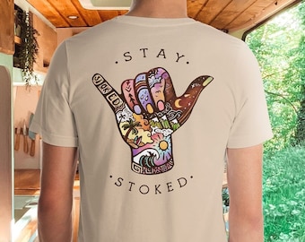 May include: A cream-coloured t-shirt with a colourful hand-shaped graphic on the back. The graphic features a beach scene, mountains, and a night sky, with the words ". STAY. STOKED." printed above and below the hand.