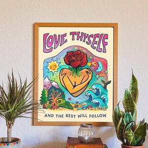 Puede incluir: Una impresión artística enmarcada con las palabras "LOVE THYSELF" y "AND THE REST WILL FOLLOW". La obra presenta una figura en forma de corazón abrazando una rosa, con un paisaje colorido de montañas, árboles y olas. El marco es de madera.