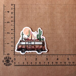 May include: A vintage camper van with a red and white tent on top, surrounded by cacti and a sunset in the background. The van is brown with a ladder on the side and the text "TB" on the back.