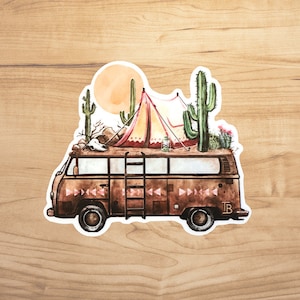 May include: A vintage brown camper van with a ladder on the side, decorated with a pink and yellow tent, a skull, and cacti on the roof. The van has a white outline and the letters "TB" on the side.