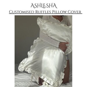 May include: A white satin pillow cover featuring ruffled edges and tie closures at the corners. The text ASHLESHA CUSTOMISED RUFFLES PILLOW COVER is displayed above the item. The fabric has a smooth, reflective finish with gathered ruffles.