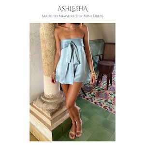 May include: A light blue silk mini dress with a bow detail. The dress is strapless and has a flowing, lightweight design. The text "ASHLESHA MADE TO MEASURE SILK MINI DRESS" is at the top of the image.