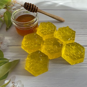 May include: Five yellow honeycomb-shaped soap bars arranged in a cluster on a white wooden surface. A glass jar of honey and a wooden honey dipper are in the background.