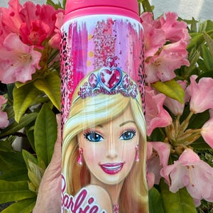 May include: A water bottle with a pink lid and a design featuring Barbie. The bottle's design includes a portrait of Barbie, a tiara, and the word "Barbie". The background has pink leopard print and glitter. The bottle is white and pink.