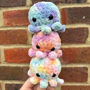 May include: Three crocheted octopus plushies stacked on top of each other. The top octopus is light blue and purple, the middle octopus is orange and pink, and the bottom octopus is yellow, orange, and blue.