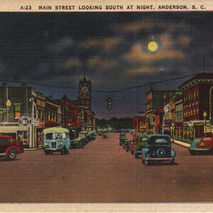 May include: A vintage postcard depicting a nighttime view of Main Street in Anderson, South Carolina. The street is lined with buildings, some with illuminated signs, and there are cars driving down the street. A full moon is visible in the sky.