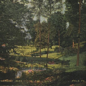 Op de afbeelding: Oude ansichtkaart met een schilderachtig uitzicht op Fairmount Park in Philadelphia, PA. De afbeelding toont een weelderig landschap met bomen, een vijver en een kleine waterval. De tekst "Fairmount Park in the Dell" is zichtbaar.