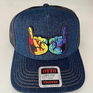 May include: A blue denim trucker hat with a rainbow-colored hand gesture patch. The hat has a mesh back and a snap closure. The label on the hat reads "OTTO FIVE STAR TRUCKER CLASSIC MESH BACK CLASSIC FIT".
