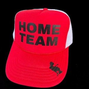 May include: A bright red trucker hat featuring a white mesh back and a solid red front panel. The text "HOME TEAM" is printed in bold black letters on the front, with a small black silhouette of a person sitting on a chair on the brim.