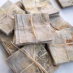 Könnte beinhalten: Eine Sammlung quadratischer Stoffservietten mit botanischen Drucken in gedeckten Erdtönen wie Beige, Braun und Grün. Jeder Stapel Servietten ist mit dünner, heller Schnur zusammengebunden und zeigt natürliche Blattmuster auf der Textiloberfläche.