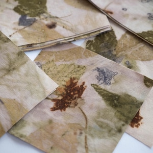 May include: A collection of square fabric napkins featuring botanical prints in shades of olive green, tan, and deep brown. The textile surface displays organic leaf and flower imprints with visible veins and textures, creating a natural pattern.