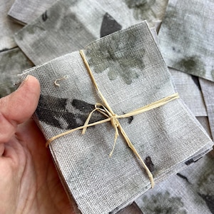 May include: A stack of square, grey linen coasters with a dark, abstract pattern. The coasters are tied together with natural twine. The coasters have a textured appearance and a neutral colour palette.