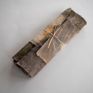 Op de afbeelding: Een bundel stofresten in tinten bruin, beige en groen, gebonden met touw. De stof heeft een ruw, getextureerd oppervlak en lijkt handgekleurd te zijn.