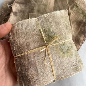 Può includere: Un mucchio di quadrati di tessuto di lino beige, alcuni con bordi sfilacciati, legati con un nastro di rafia naturale. Il tessuto presenta sottili sfumature marroni e verdi, suggerendo un processo di tintura naturale. Ulteriori pezzi di tessuto sono sparsi.