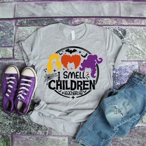 May include: A gray t-shirt with a black circle design featuring three witches with the text "I smell children #teacherlife".