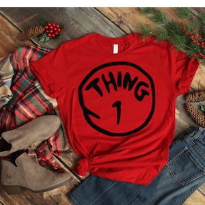 May include: Red t-shirt with a black circle graphic that says "Thing 1" in white letters. The shirt is folded and laid on a wooden surface with a plaid blanket, brown boots, and blue jeans.