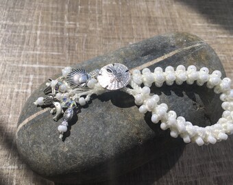 May include: White beaded bracelet with a silver charm featuring a seashell and a sand dollar. The bracelet is on a gray rock.