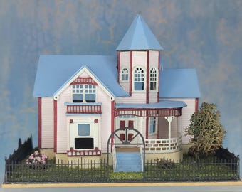 May include: A miniature Victorian-style dollhouse with a pink exterior, blue roof, and a small tower. The house features a porch, windows, and a black fence. A small tree and pink flowers are also present.