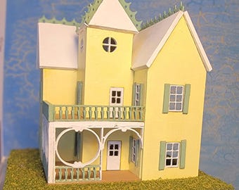 May include: A miniature yellow house with white trim, green accents, and a green roof. The house has a porch, balcony, and multiple windows. It sits on a base of artificial grass, creating a charming, detailed model.