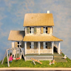 May include: A miniature two-story house with a porch, shutters, and a textured roof. A swing set, seesaw, and small figures of a person, cat, and bird are on the grassy base. The house is painted in neutral tones with green accents.