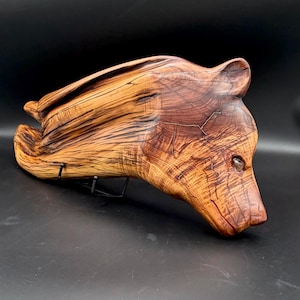 May include: A wooden bear sculpture with a natural wood grain finish. The bear is depicted in a reclining pose, with detailed carving of the head and body. The sculpture is mounted on a black metal stand.