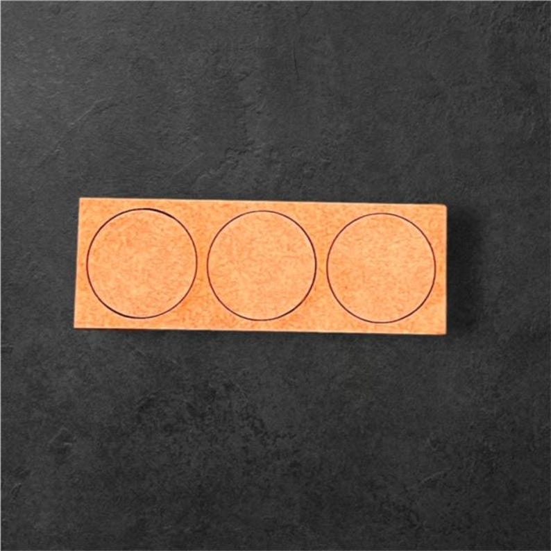 May include: A rectangular, light orange coaster with three circular cutouts. The coaster is made of a textured material and has black outlines around each circle. The background is a dark gray, textured surface.