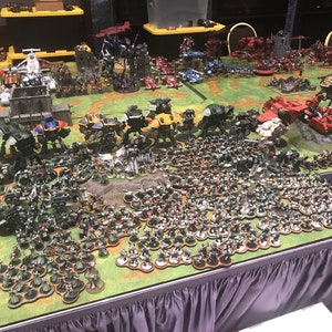 May include: A large tabletop wargaming scene with a green felt playing surface.  The scene features dozens of miniature figures, including several large robots, tanks, and infantry.  The figures are painted in various colours, including green, red, and yellow.