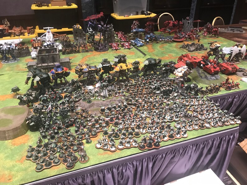 May include: A large tabletop wargaming scene with a green felt playing surface.  The scene features dozens of miniature figures, including several large robots, tanks, and infantry.  The figures are painted in various colours, including green, red, and yellow.