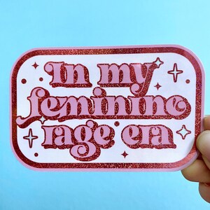 May include: Pink and white sticker with glitter and the text "In my feminine rage era".