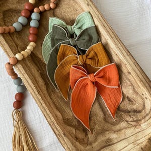 May include: Four fabric hair bows in a wooden bowl. The bows are in shades of green, brown, orange, and red. Each bow has a white trim.