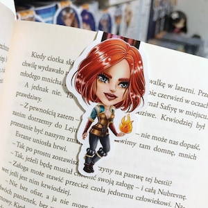 Peut inclure: Gros plan d'un marque-page illustrant un dessin animé d'une femme aux cheveux roux, portant une veste en cuir marron et tenant une torche enflammée. Le marque-page est placé sur une page d'un livre avec du texte en polonais.