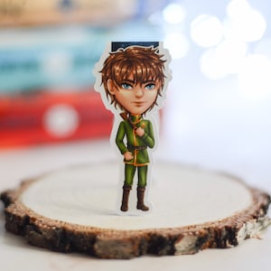 Può includere: Un segnalibro con un personaggio dei cartoni animati, un giovane con i capelli castani, che indossa un'uniforme militare verde e stivali. Il segnalibro è in piedi su una fetta di legno.